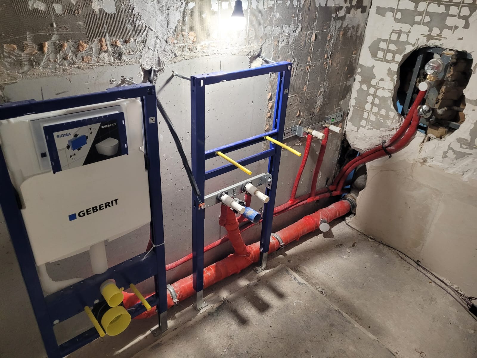 Stelaż podtynkowy Geberit Sigma w trakcie montażu, widoczne rury kanalizacyjne w kolorze czerwonym i instalacja wodna w surowym stanie, ściany w trakcie remontu, widoczne ślady po skuciu płytek.