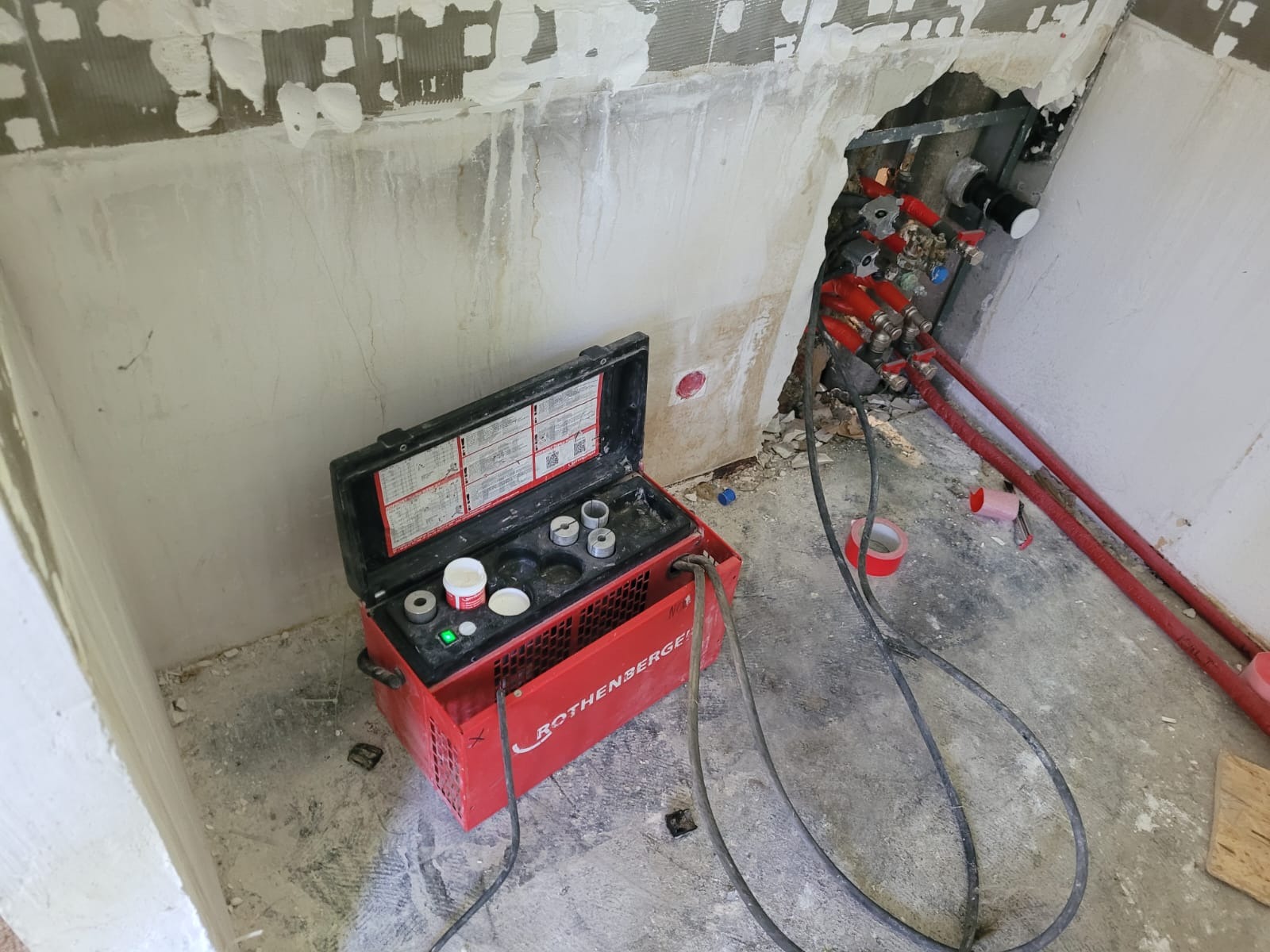 Otwarta walizka narzędziowa Rothenberger, z widocznymi elementami do zaciskania rur, stoi na podłodze w trakcie prac hydraulicznych; w tle ściana z resztkami tynku i widoczne rury instalacji...