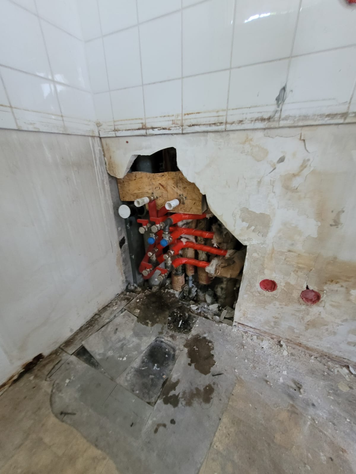 Odsłonięte rury instalacji wodnej w ścianie podczas remontu łazienki, widoczne czerwone rury z niebieskimi zaworami, drewniane elementy konstrukcyjne i fragmenty starej instalacji.