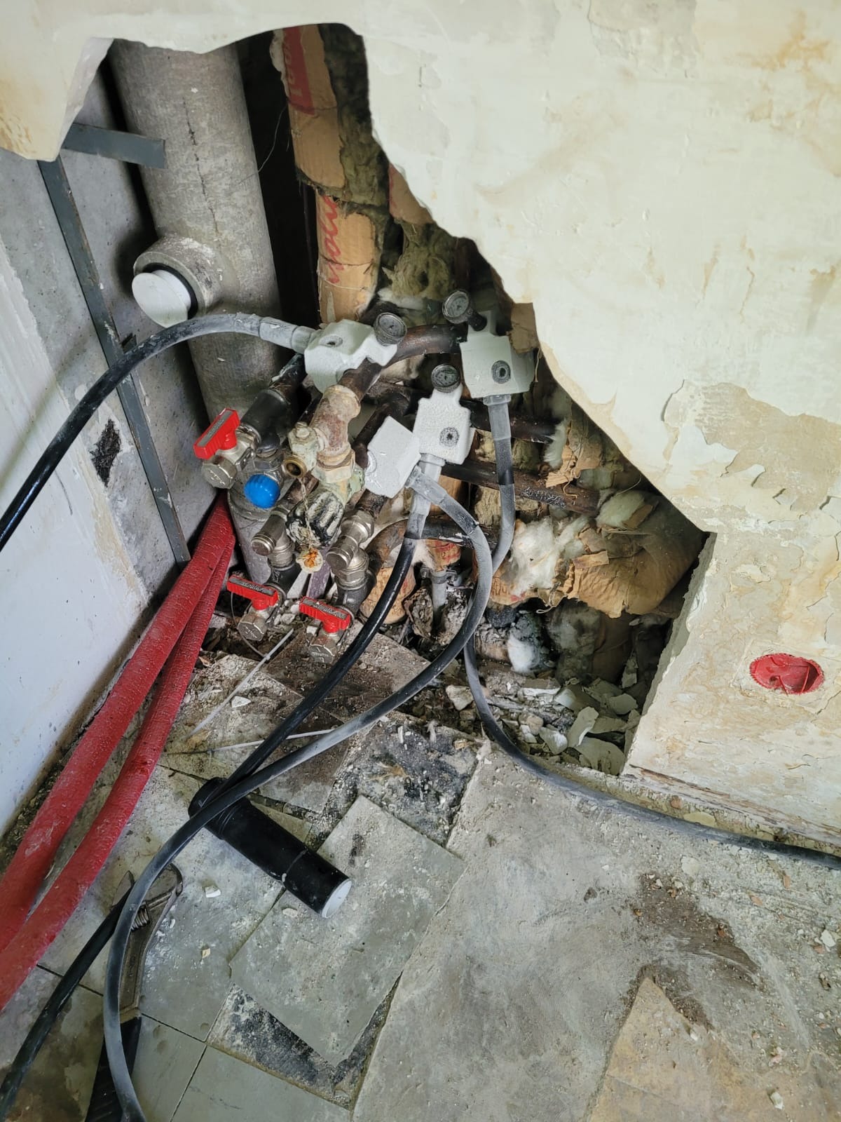 Odsłonięta instalacja hydrauliczna w ścianie z widocznymi rurami, zaworami z czerwonymi i niebieskimi pokrętłami, przewodami elektrycznymi, fragmentami starego tynku i izolacji.