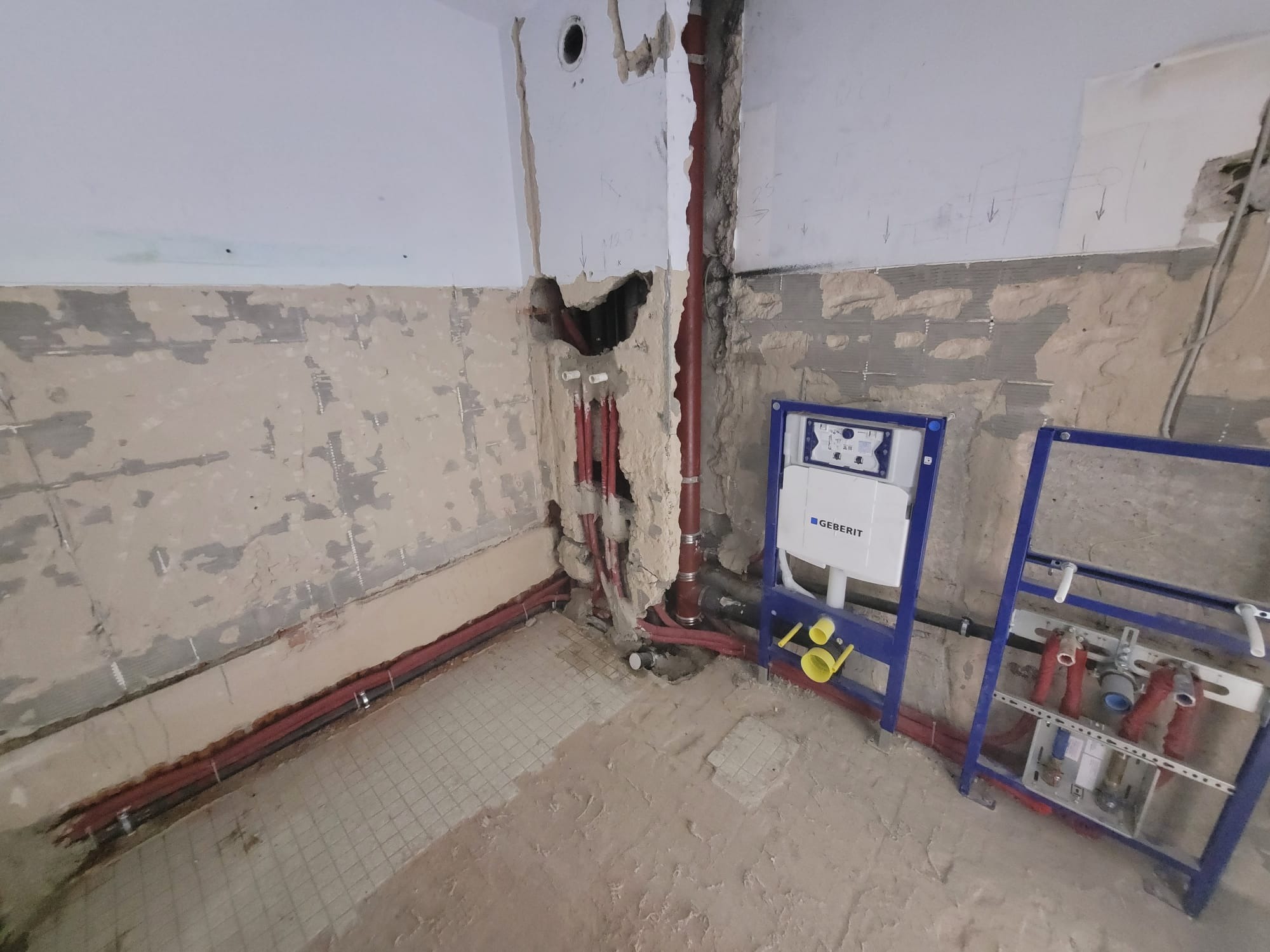 Wnętrze łazienki w trakcie przeróbki instalacji hydraulicznej z widocznym stelażem podtynkowym Geberit, nowymi rurami w kolorze czerwonym i fragmentami starej instalacji oraz śladami po kuciu...