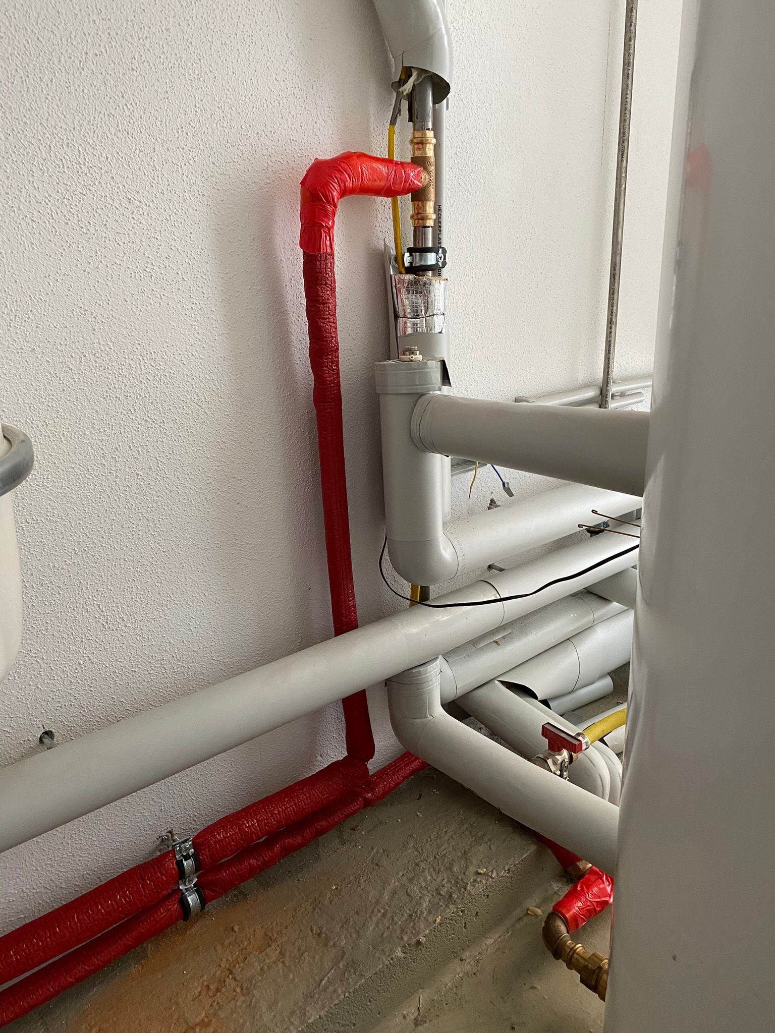 Szare rury instalacyjne z czerwoną izolacją termiczną, fragment bojlera, biała ściana o chropowatej fakturze i betonowa podłoga.