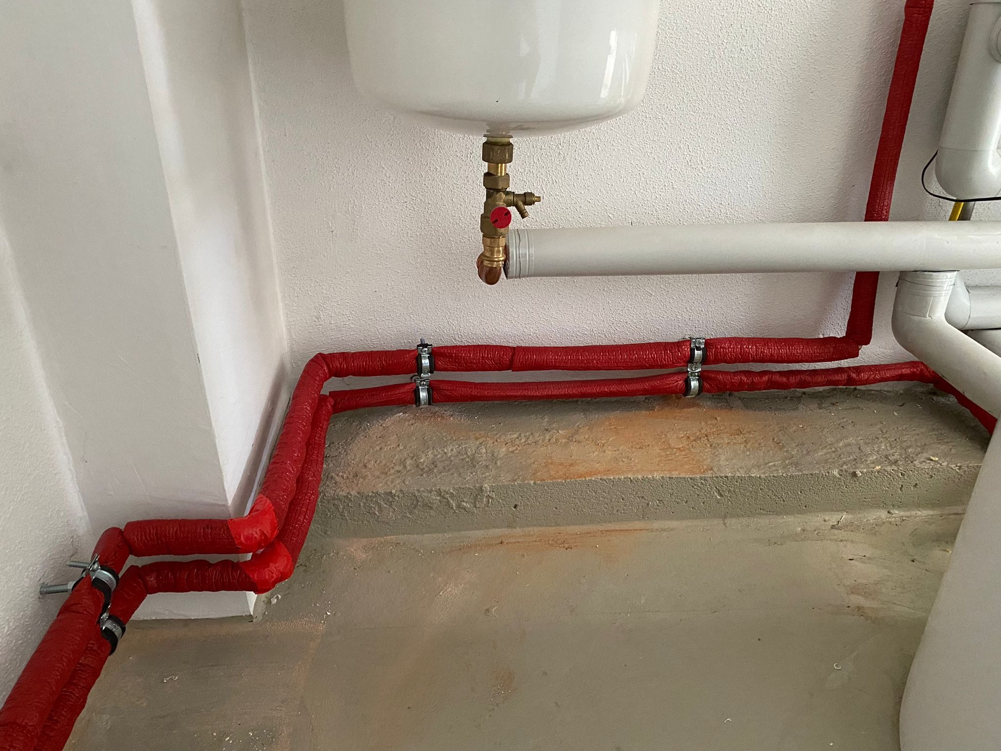 Czerwone rury grzewcze z izolacją termiczną, mocowane do ściany i podłogi, widoczny zawór i fragment instalacji pod umywalką.