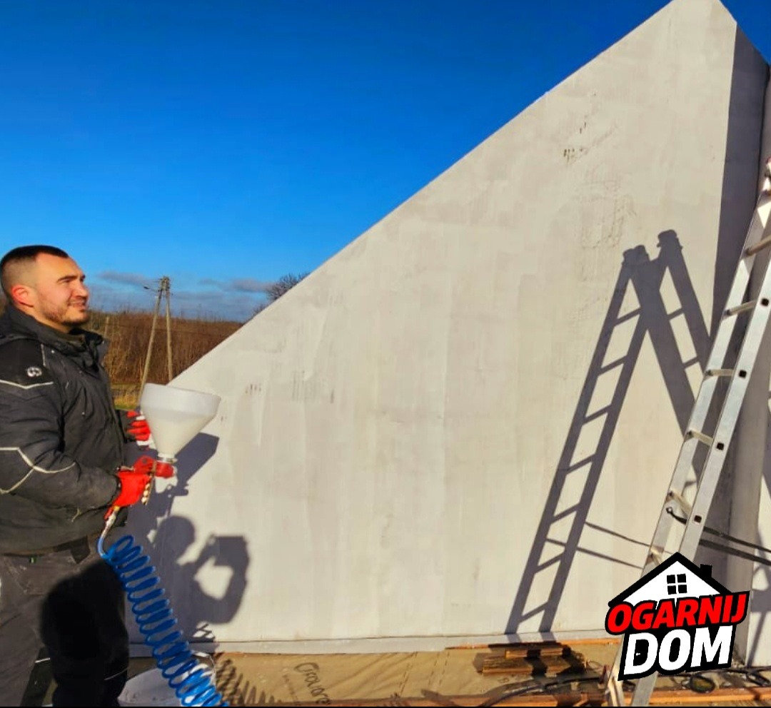 Pracownik w czerwonych rękawicach nanosi tynk strukturalny na ścianę domu za pomocą pistoletu natryskowego; widoczna drabina oparta o ścianę, błękitne niebo w tle.