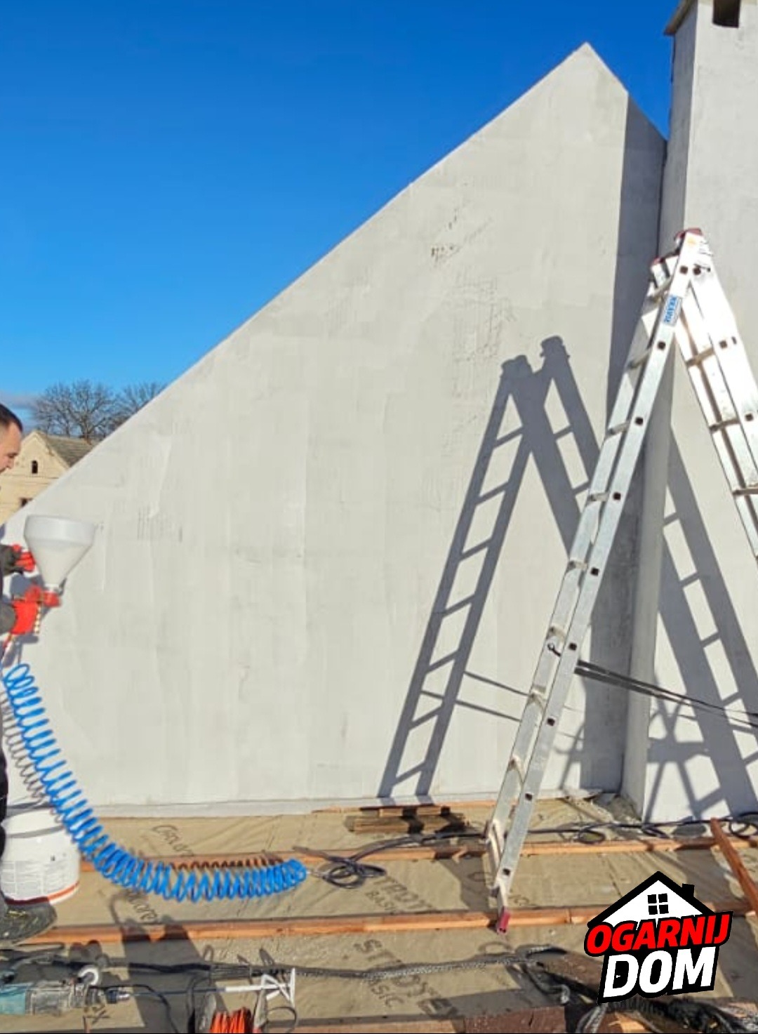 Nakładanie tynku maszynowego na elewację domu jednorodzinnego przy użyciu agregatu tynkarskiego, widoczna drabina i niebieski przewód sprężonego powietrza.
