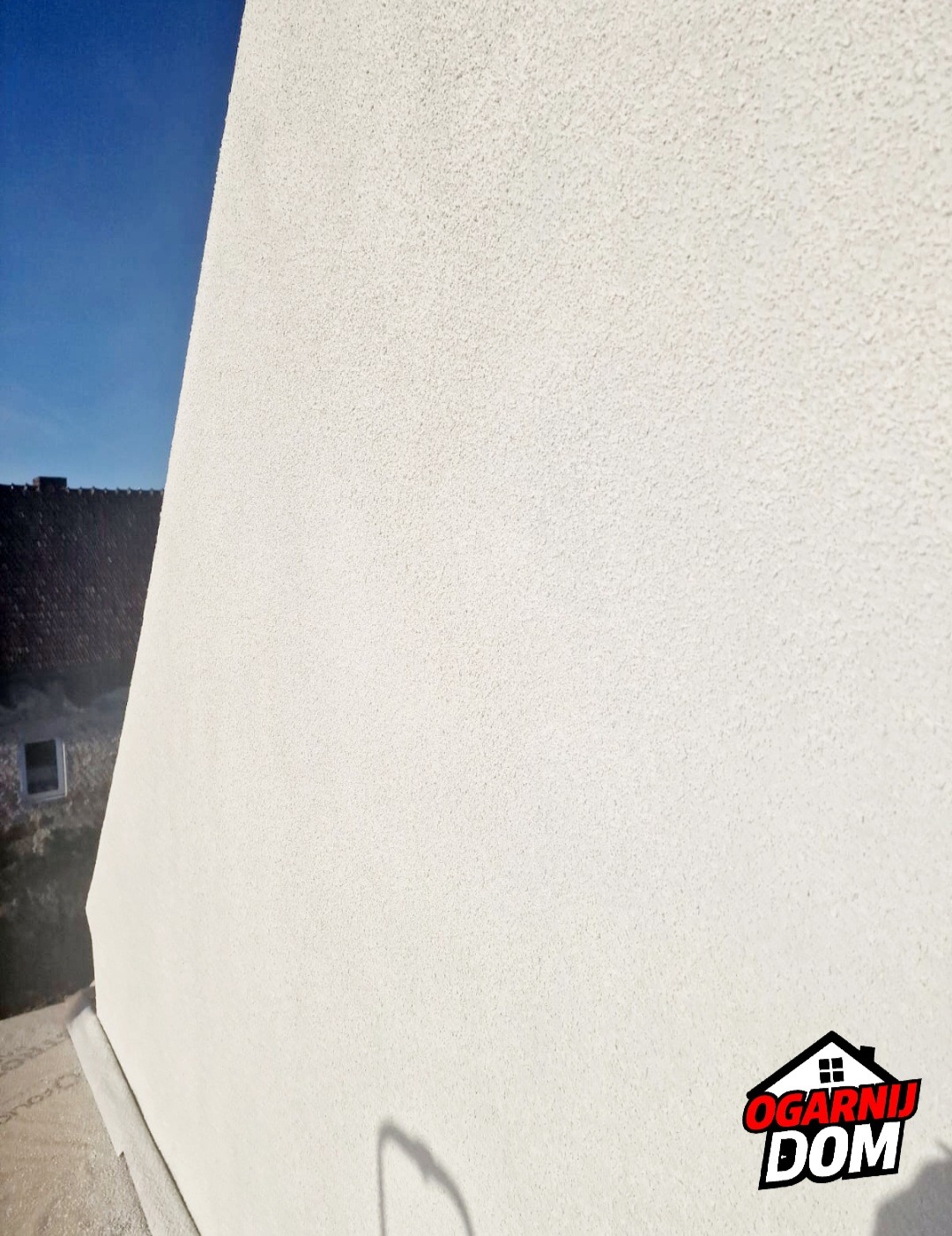 Fragment ściany budynku z nowym, jasnym tynkiem strukturalnym, widoczny cień i logo firmy w prawym dolnym rogu, w tle fragment dachu i okno.