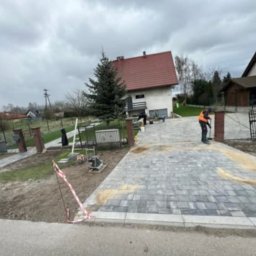Adi-bruk - Układanie szarej kostki brukowej na podjeździe przed domem z czerwoną dachówką; pracownik w pomarańczowej kamizelce zagęszcza piasek fugowy, widoczne narzędzia i maszyny brukarskie, częściowo...