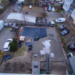 Adi-bruk - Widok z góry na plac budowy z pracownikami układającymi czarną nawierzchnię, prawdopodobnie pod kostkę, w pobliżu parkingu i budynku mieszkalnego.