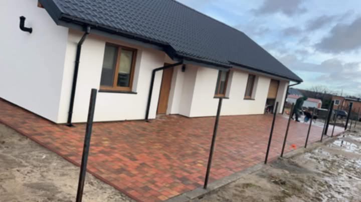 Nowoczesny dom z wyłożonym kostką brukową podjazdem w odcieniach czerwieni i brązu, widoczne słupki ogrodzeniowe i pracownik wykonujący prace wykończeniowe.