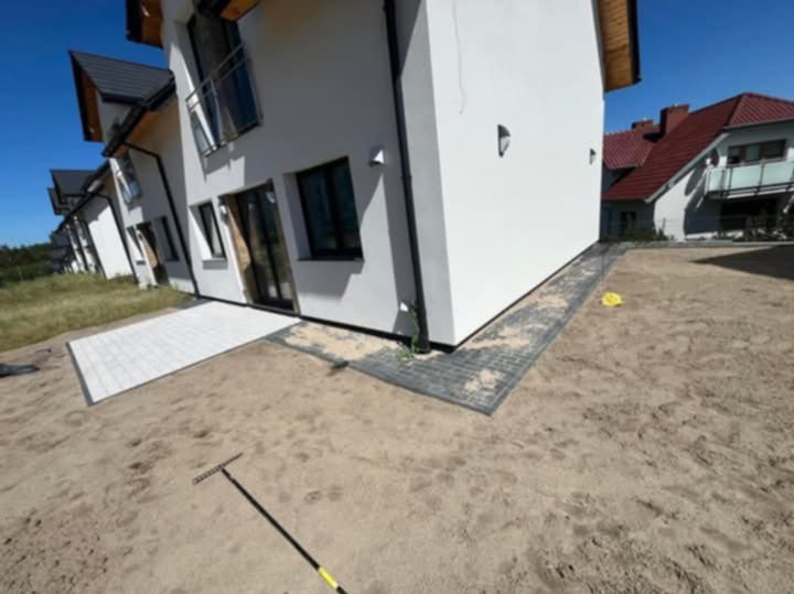 Nowoczesny dom z białymi ścianami i częściowo ułożonym chodnikiem z szarej kostki brukowej wokół budynku, widoczna grządka z piaskiem i grabie ogrodowe.