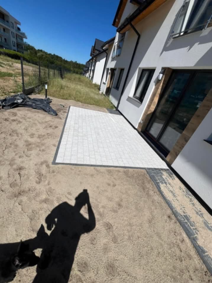 Biała kostka ułożona przy szeregówce, widoczny cień osoby robiącej zdjęcie i fragment psa, teren przygotowany pod dalsze prace.