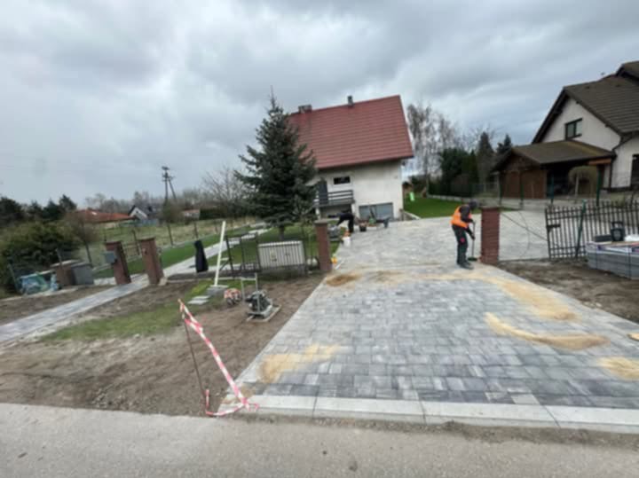 Układanie szarej kostki brukowej na podjeździe przed domem z czerwoną dachówką; pracownik w pomarańczowej kamizelce zagęszcza piasek fugowy, widoczne narzędzia i maszyny brukarskie, częściowo...