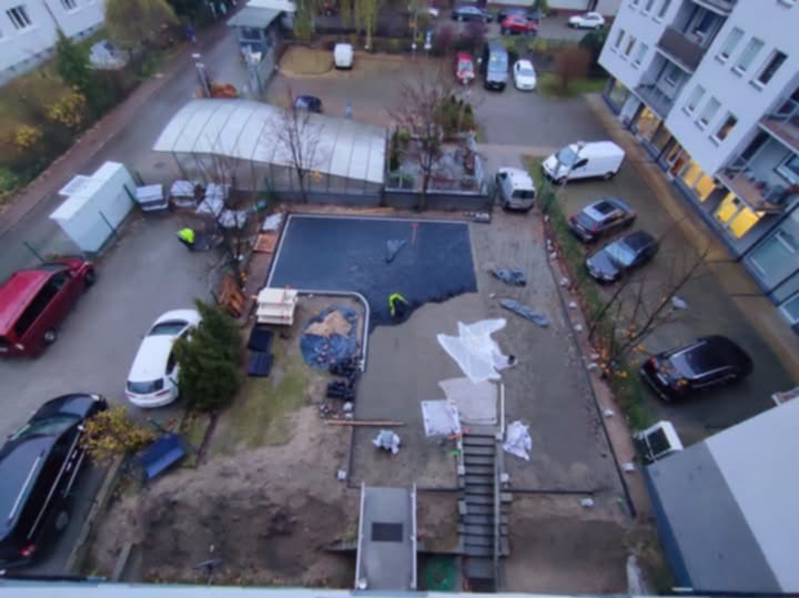 Widok z góry na plac budowy z pracownikami układającymi czarną nawierzchnię, prawdopodobnie pod kostkę, w pobliżu parkingu i budynku mieszkalnego.