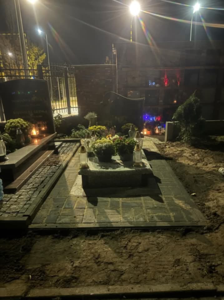 Cmentarz nocą: wybrukowany grób z kwiatami i zniczami, oświetlony latarniami ulicznymi, z widoczną świeżo usypaną ziemią obok.