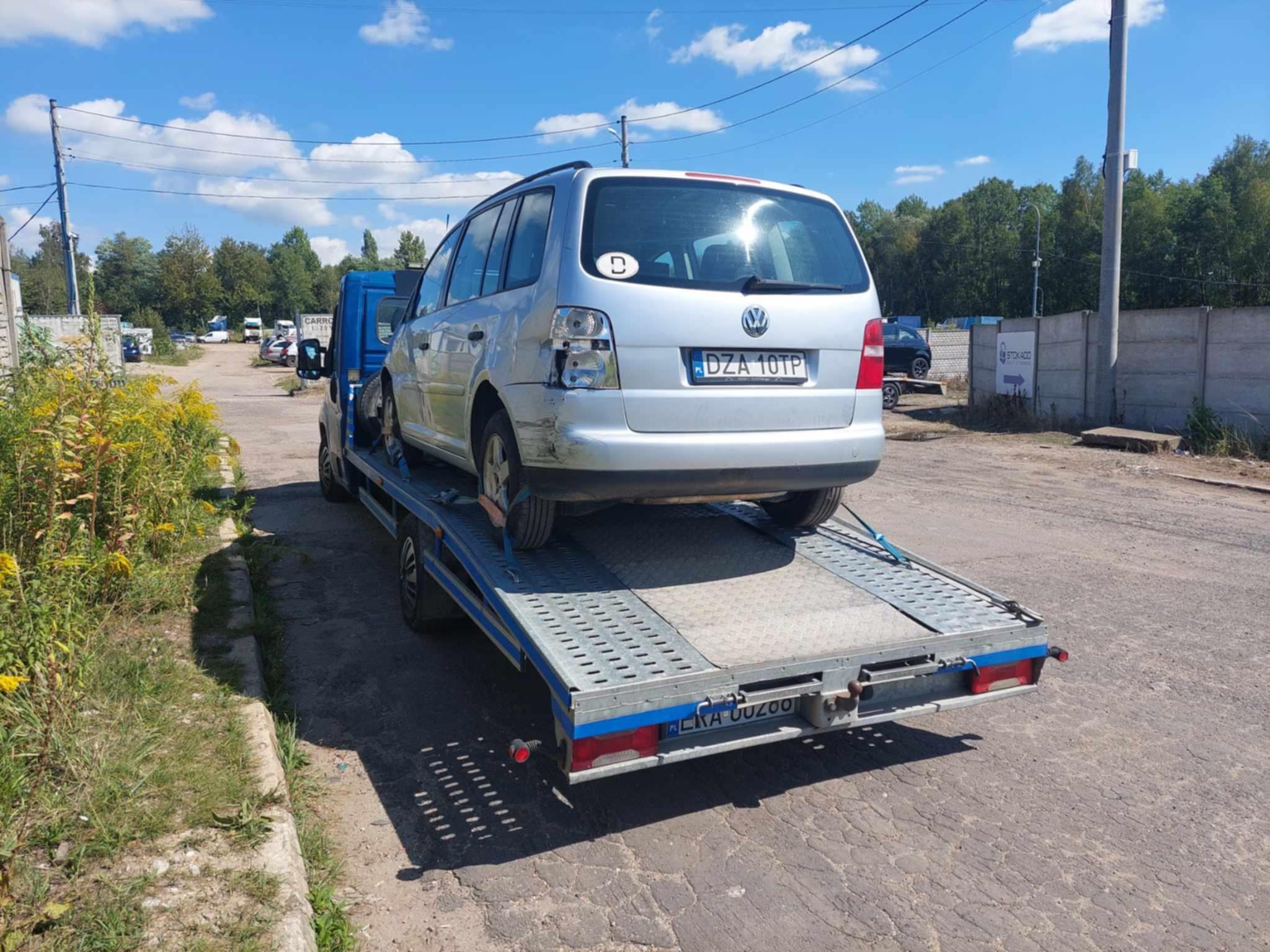 Srebrny samochód osobowy Volkswagen Touran z uszkodzonym tyłem, z niemiecką naklejką i tablicą rejestracyjną, załadowany na niebieską lawetę na zewnątrz w słoneczny dzień.