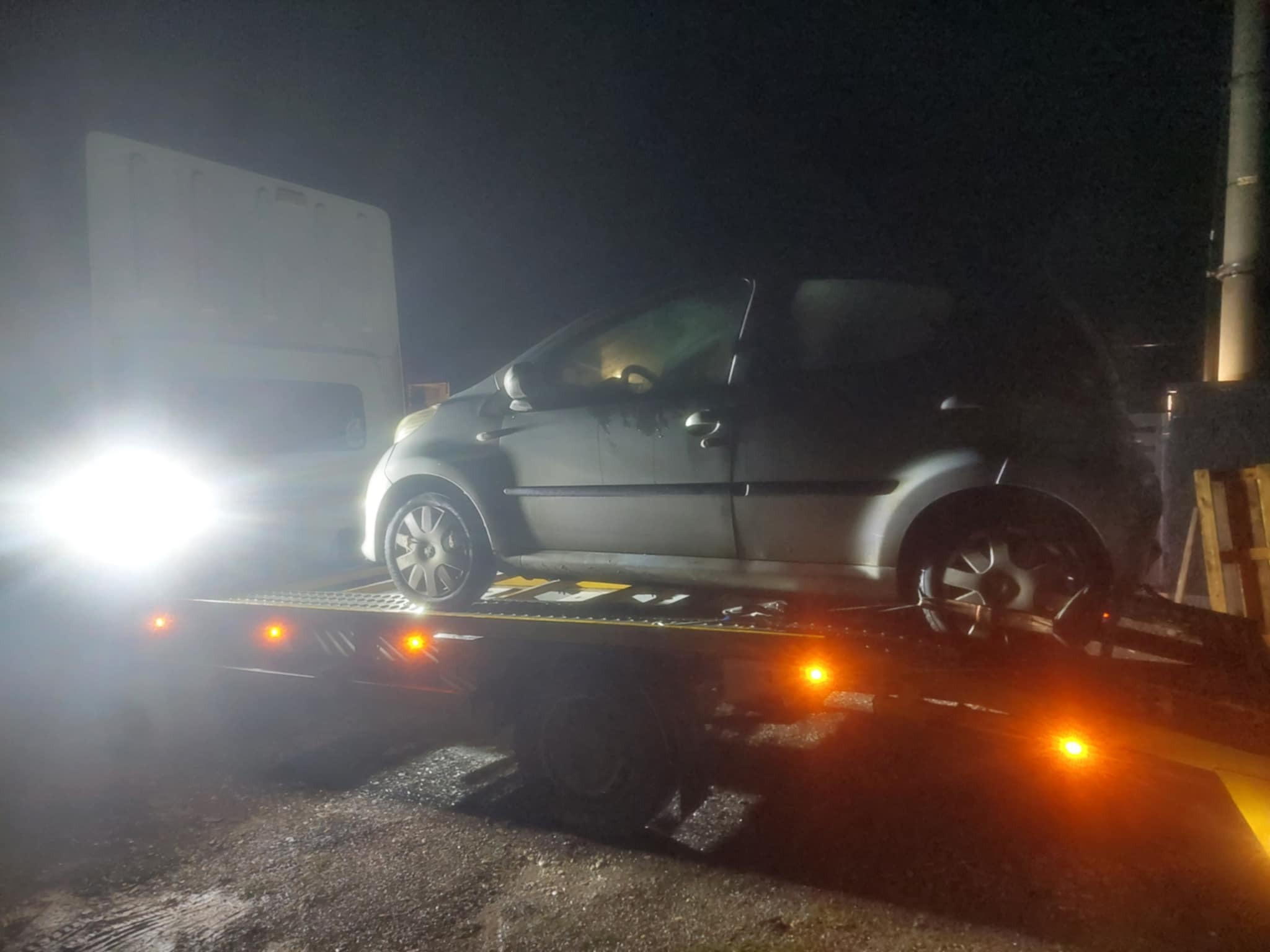 Samochód osobowy srebrny załadowany na lawetę w nocy, widoczny biały van w tle, oświetlenie nocne.