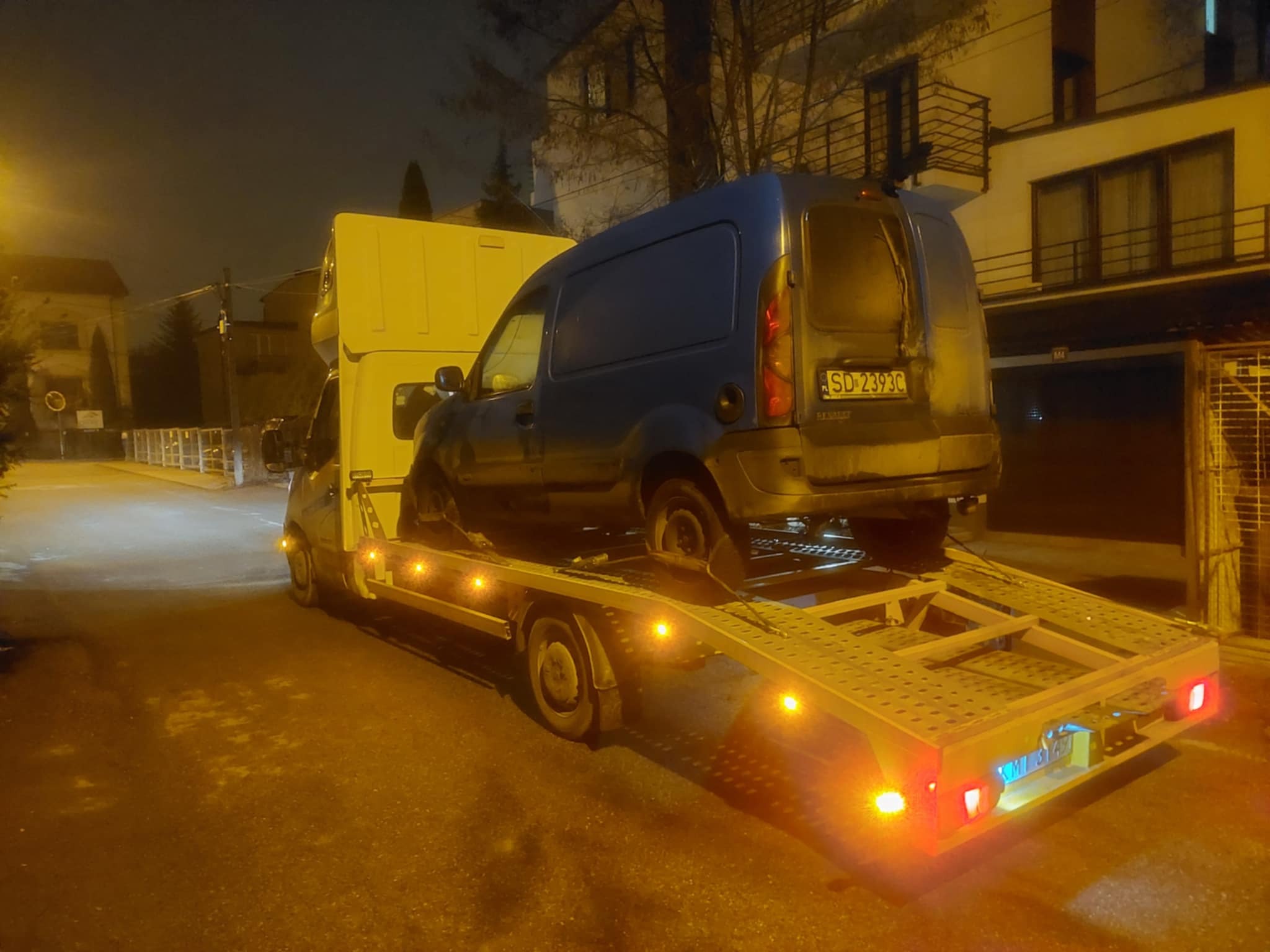 Niebieski samochód dostawczy Renault Kangoo z uszkodzonym kołem, przewożony na żółtej lawecie w nocy, oświetlony światłami ulicznymi. Widoczny numer rejestracyjny pojazdu.