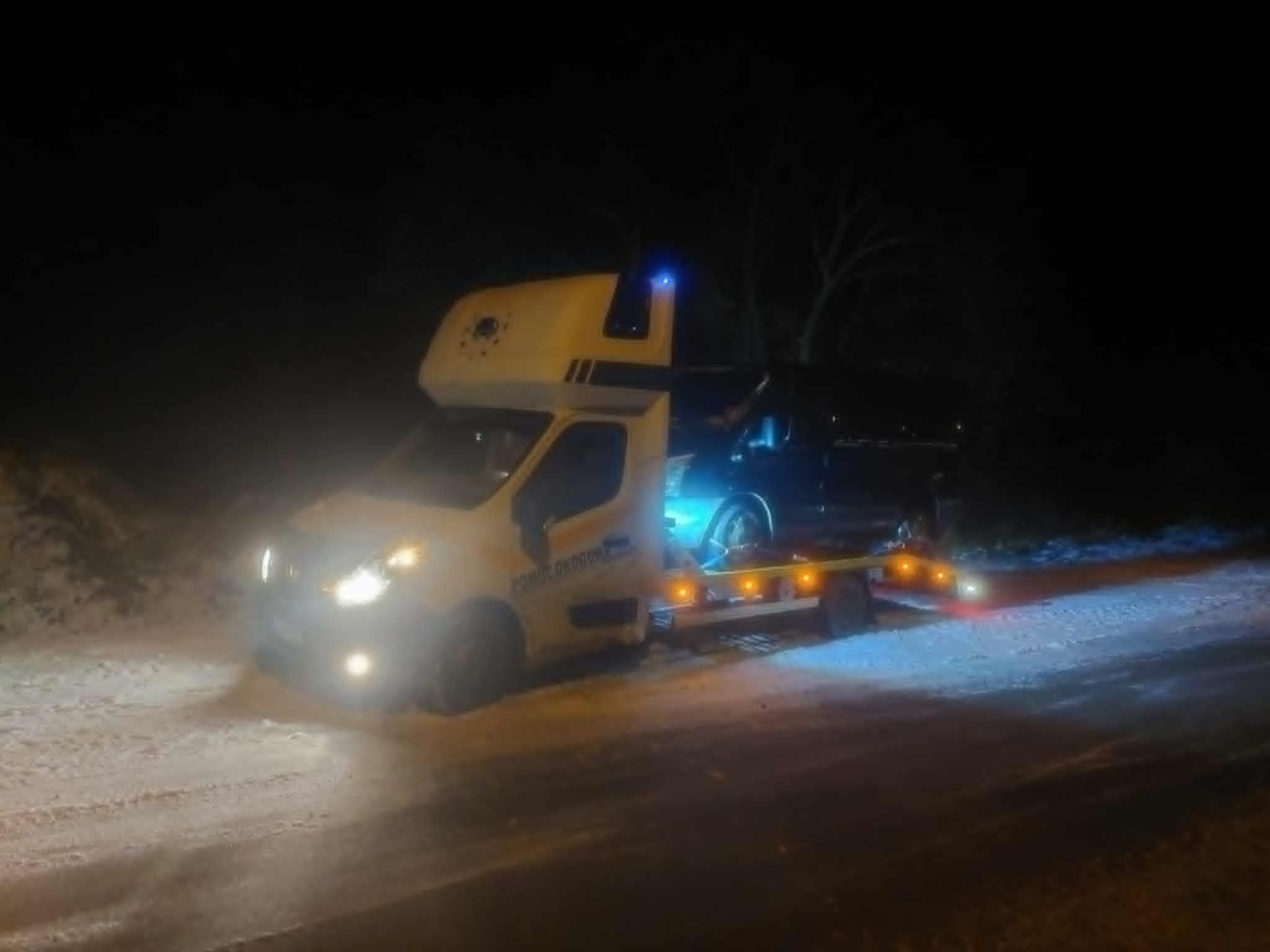 Żółta laweta z niebieskim kogutem przewozi czarny samochód na ośnieżonej drodze nocą.