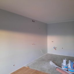 Fixbud Pro - Pokój w trakcie malowania na szaro, widoczne puszki z farbą, szpachla na ścianie i przygotowane podłoże pod panele.