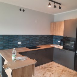 Fixbud Pro - Nowoczesna kuchnia w trakcie montażu: szare szafki dolne i górne, drewniane blaty, niebieska mozaika na ścianie, marmurowa podłoga, karton z elementami montażowymi i butelka wody na blacie wyspy.