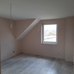 Fixbud Pro - Pomalowane na jasnoróżowo poddasze z oknem, szarymi panelami podłogowymi i wiszącymi przewodami elektrycznymi bez lamp.