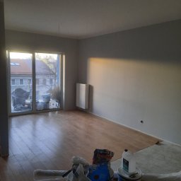 Fixbud Pro - Pokój w trakcie odświeżania: pomalowane na szaro ściany, częściowo ułożone panele podłogowe, widoczne puszki z farbą i narzędzia malarskie przy balkonie.