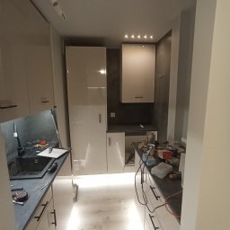 Fixbud Pro - Kuchnia w trakcie remontu: widoczne białe szafki z czarnymi uchwytami, czarny blat z narzędziami, podświetlenie LED pod szafkami, sufit z oświetleniem punktowym i narzędzia na blacie.