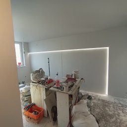 Fixbud Pro - Wnętrze w trakcie prac wykończeniowych: świeżo pomalowane szare ściany z zamontowanym oświetleniem LED wzdłuż sufitu i pionowo w rogu, na podłodze widoczne ślady gładzi i farby, prowizoryczny stół...