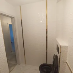 Nowoczesna łazienka z białymi, trójwymiarowymi płytkami i złotymi akcentami, czarna toaleta w minimalistycznym stylu, marmurowa podłoga, widok na otwarte drzwi do innego pomieszczenia.