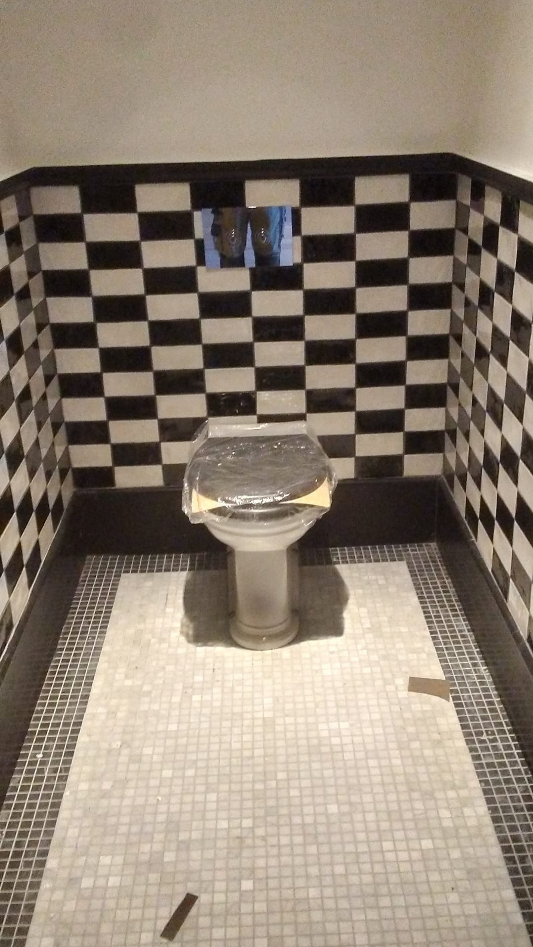 Biała toaleta z plastikową osłoną na desce, umieszczona w łazience z czarno-białą szachownicą na ścianach i mozaiką na podłodze, widoczne elementy po remoncie.