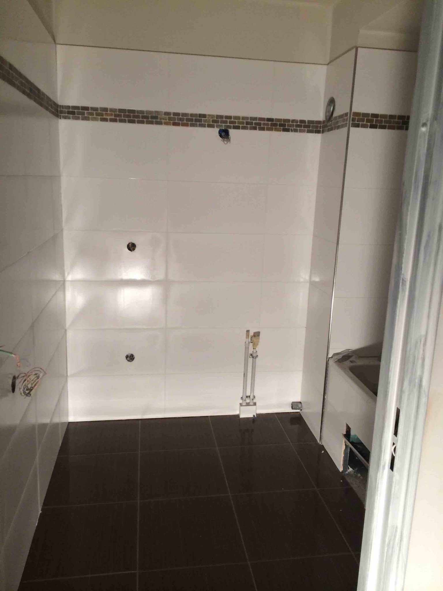 Łazienka w trakcie remontu: białe płytki ścienne z dekoracyjnym pasem mozaiki, ciemnobrązowe płytki podłogowe, widoczne rury instalacyjne i otwory na oświetlenie, częściowo zasłonięta wanna...