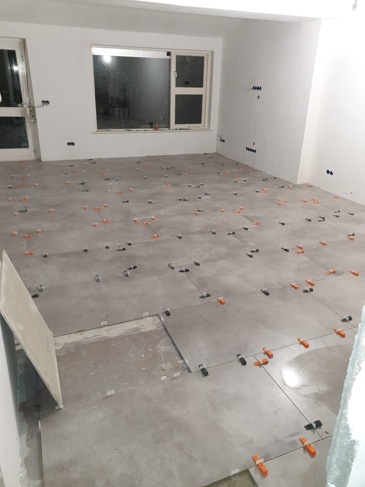 Szare płytki podłogowe w trakcie układania, z pomarańczowymi i czarnymi klipsami poziomującymi, widoczne niedokończone fragmenty podłogi i surowe ściany z wystającymi przewodami elektrycznymi.