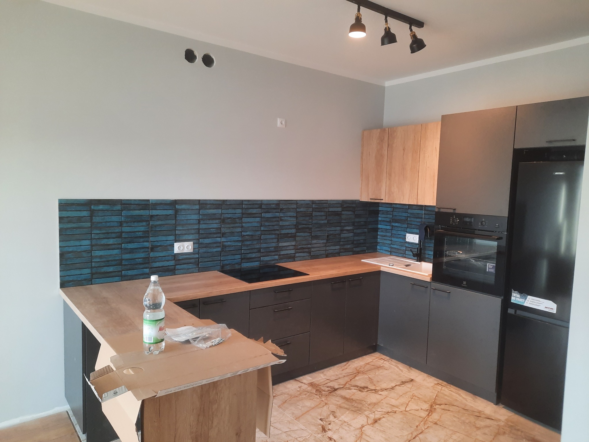 Nowoczesna kuchnia w trakcie montażu: szare szafki dolne i górne, drewniane blaty, niebieska mozaika na ścianie, marmurowa podłoga, karton z elementami montażowymi i butelka wody na blacie wyspy.