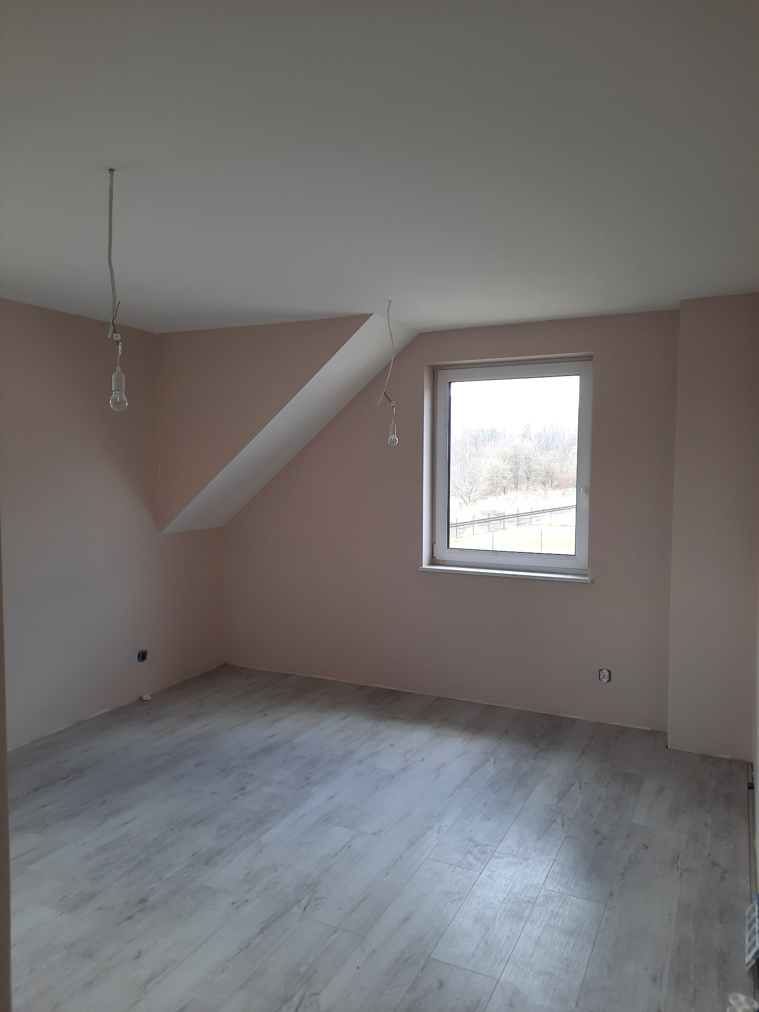 Pomalowane na jasnoróżowo poddasze z oknem, szarymi panelami podłogowymi i wiszącymi przewodami elektrycznymi bez lamp.