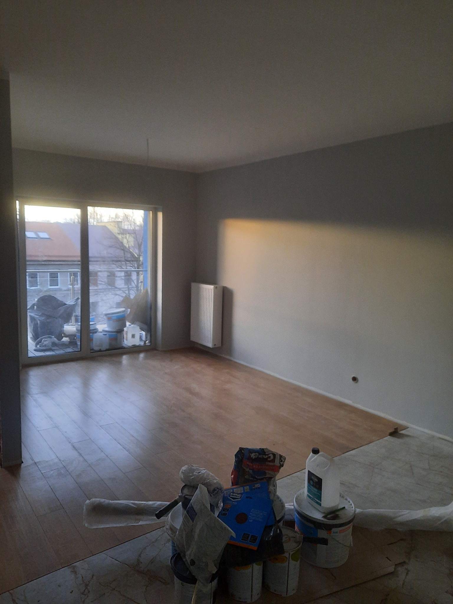 Pokój w trakcie odświeżania: pomalowane na szaro ściany, częściowo ułożone panele podłogowe, widoczne puszki z farbą i narzędzia malarskie przy balkonie.