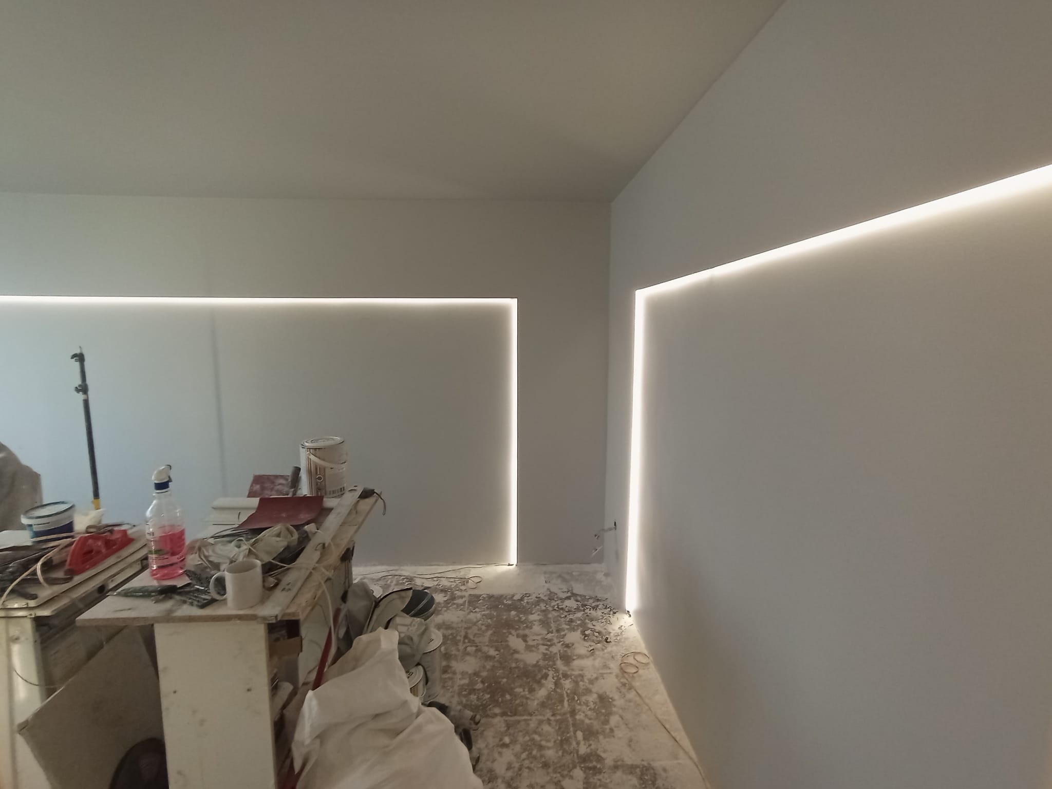 Pokój w trakcie remontu z zamontowanym oświetleniem LED wzdłuż ścian, widoczne narzędzia i materiały budowlane na prowizorycznym stole roboczym, podłoga częściowo zakryta folią malarską.