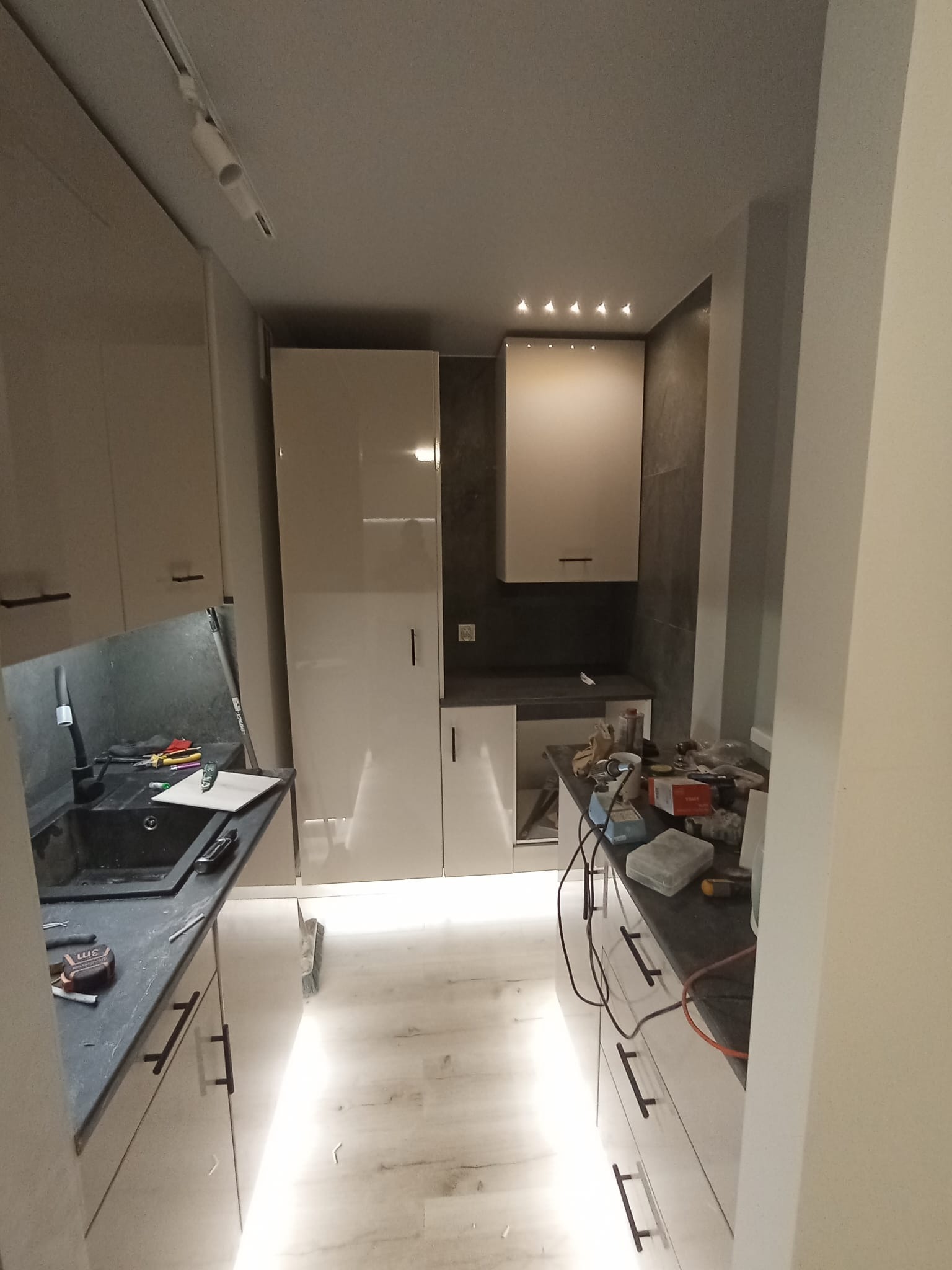 Wnętrze kuchni w trakcie prac wykończeniowych, widoczne białe, lakierowane szafki z czarnymi uchwytami, czarny zlew z baterią, narzędzia na blacie, podświetlenie LED pod szafkami i lampy sufitowe.