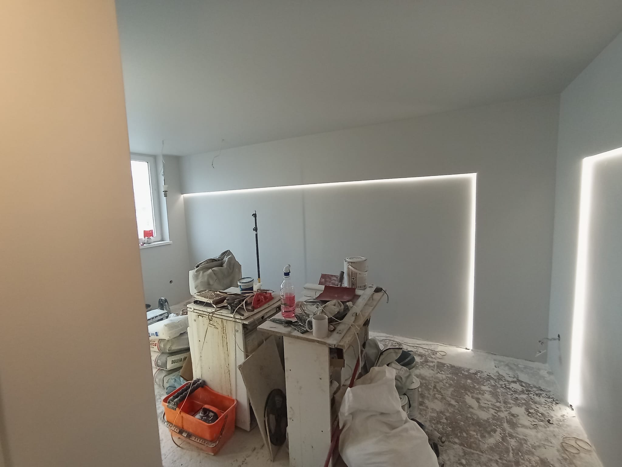 Wnętrze w trakcie prac wykończeniowych: świeżo pomalowane szare ściany z zamontowanym oświetleniem LED wzdłuż sufitu i pionowo w rogu, na podłodze widoczne ślady gładzi i farby, prowizoryczny stół...