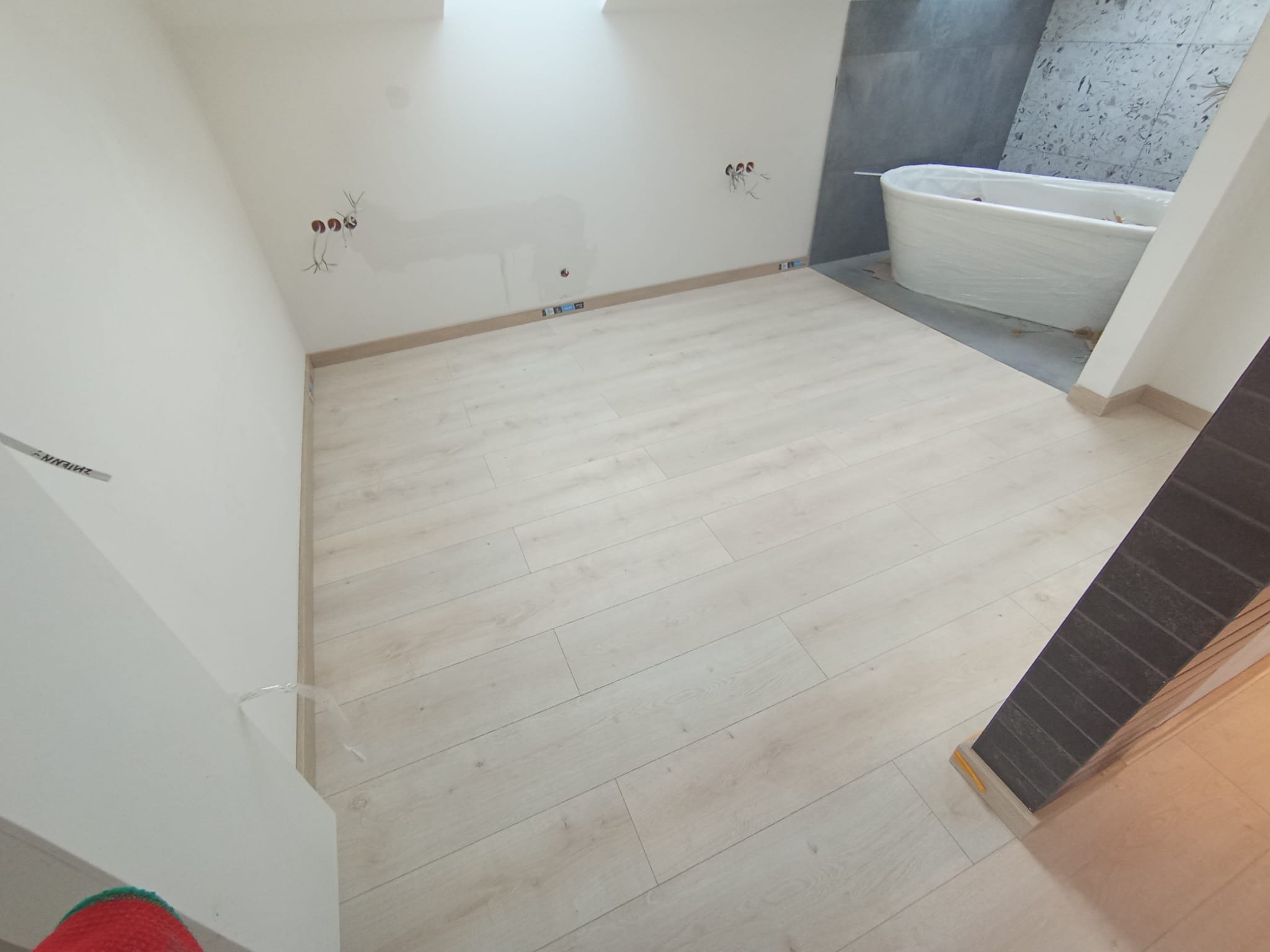 Jasne panele podłogowe ułożone w łazience, z widocznymi przygotowaniami do instalacji elektrycznej na ścianie i wanną zabezpieczoną folią.