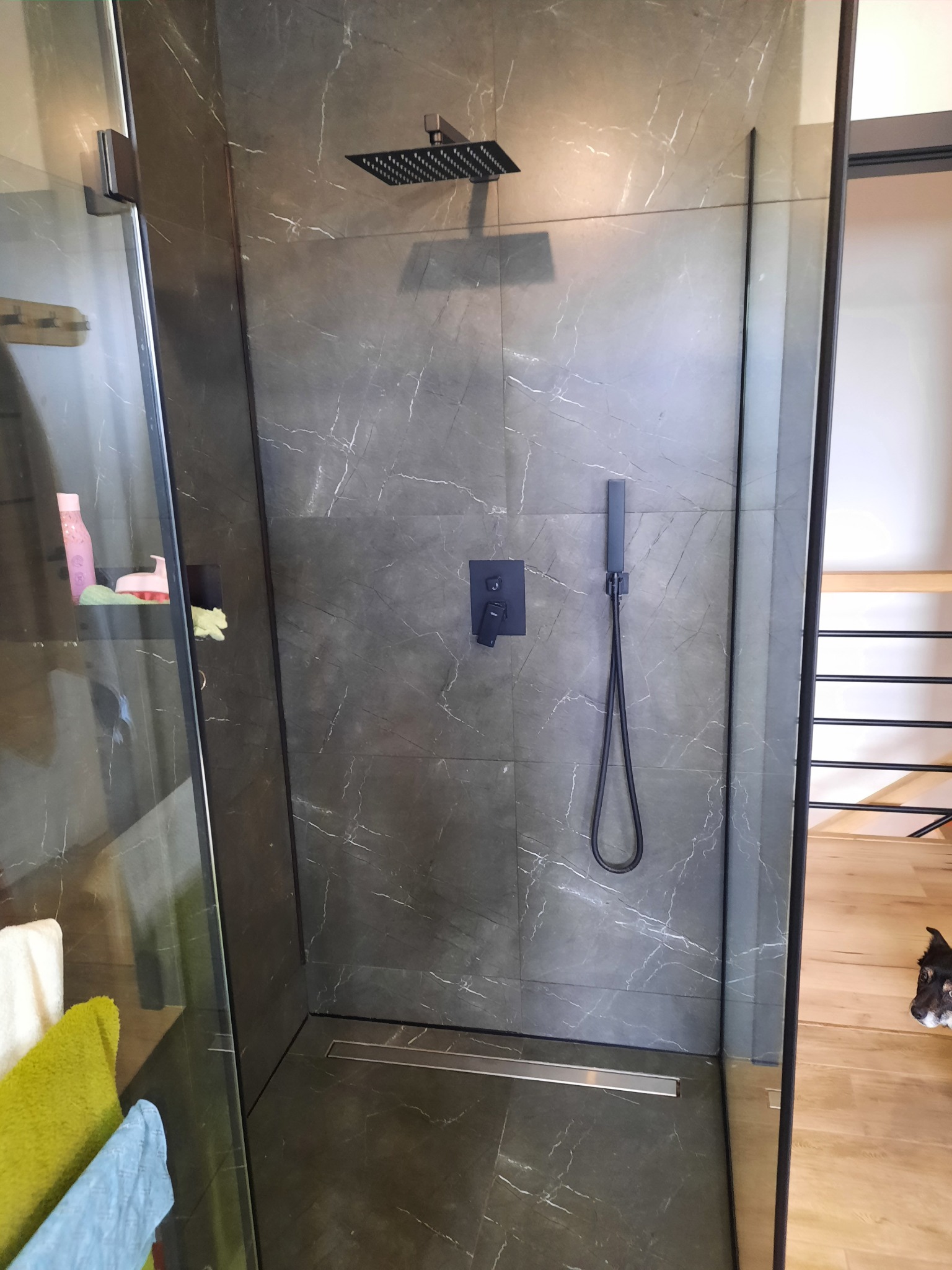 Nowoczesna kabina prysznicowa z szarymi płytkami imitującymi marmur, czarną armaturą i odpływem liniowym, widok z zewnątrz kabiny, fragment podłogi drewnianej i głowa psa w prawym dolnym rogu.