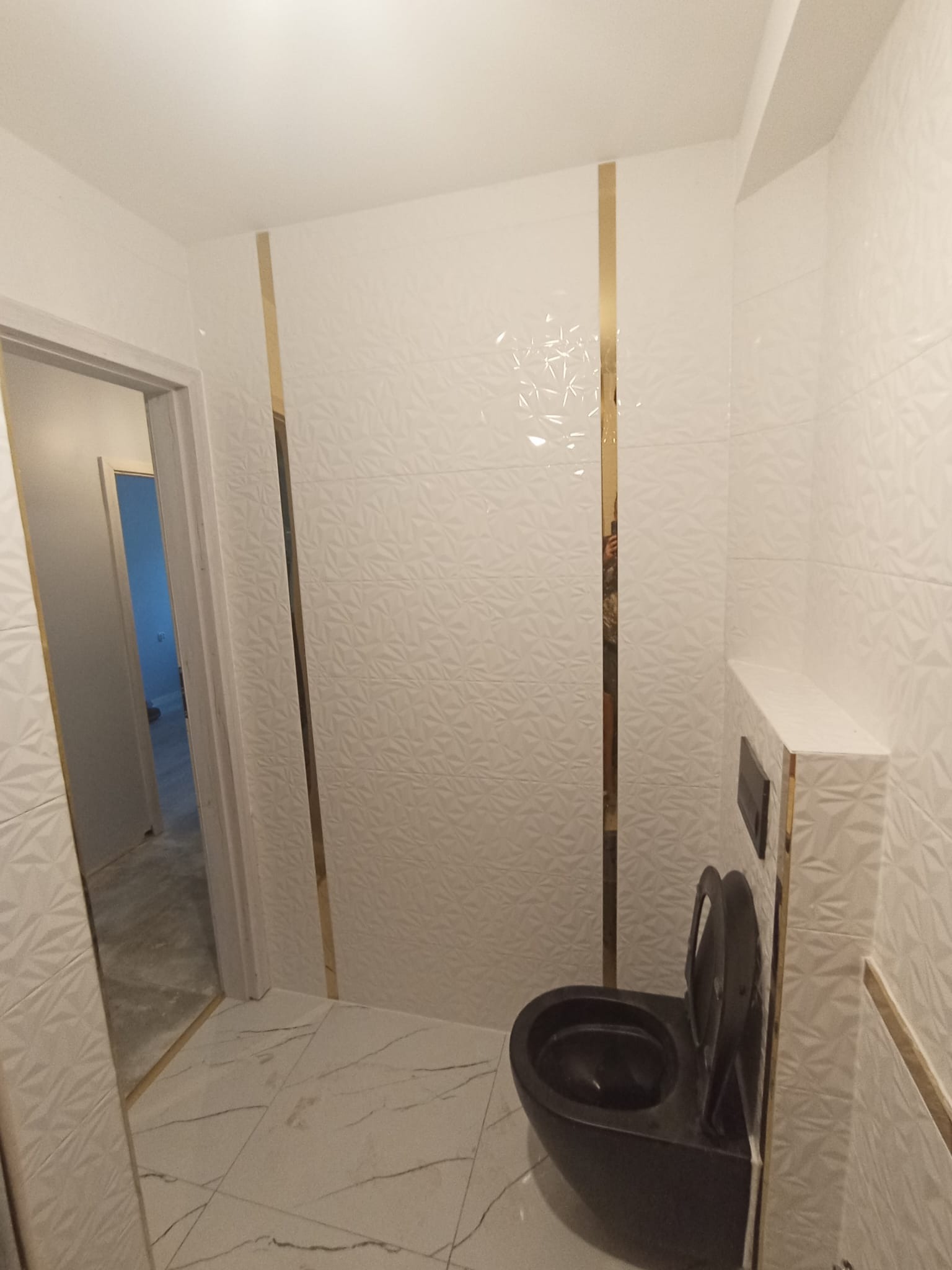 Nowoczesna łazienka z białymi, trójwymiarowymi płytkami i złotymi akcentami, czarna toaleta w minimalistycznym stylu, marmurowa podłoga, widok na otwarte drzwi do innego pomieszczenia.