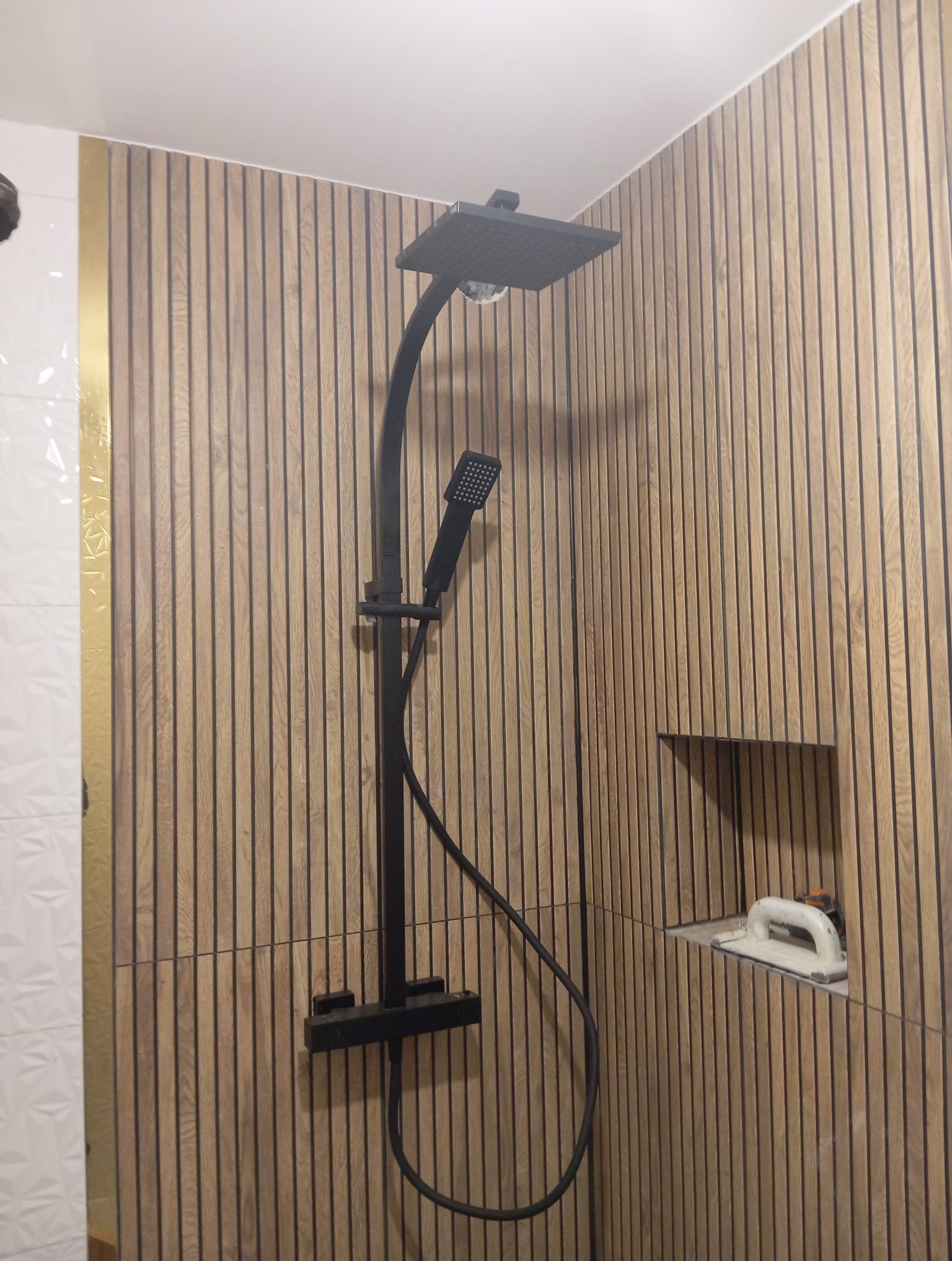 Nowoczesna kabina prysznicowa z deszczownicą i słuchawką w kolorze czarnym, na tle ściany wykończonej panelami imitującymi drewno, z widoczną wnęką i narzędziem do fugowania.