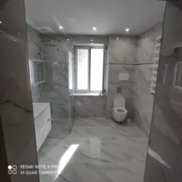 Nowoczesna łazienka wykończona płytkami imitującymi marmur, z kabiną prysznicową typu walk-in, podwieszaną toaletą i grzejnikiem drabinkowym, oświetlona punktowymi lampami sufitowymi i naturalnym...