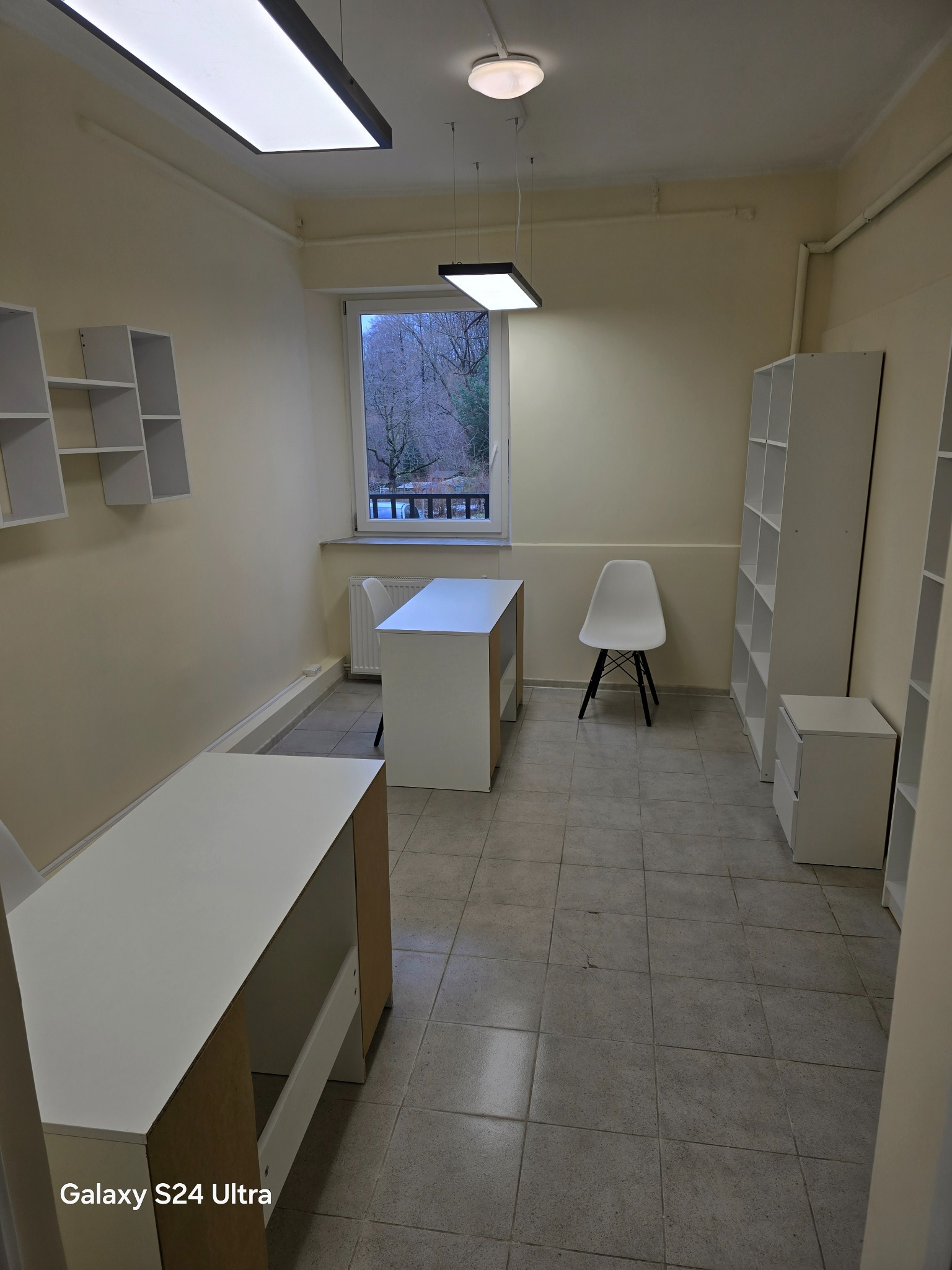 Wnętrze biura w trakcie urządzania: białe biurka z surowymi bokami, regały na ścianach, szara podłoga z płytek, widok z okna na drzewa.