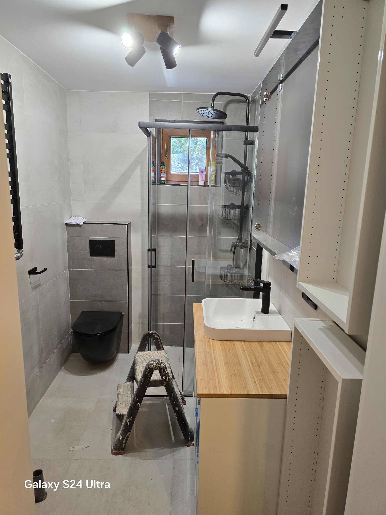 Wnętrze łazienki w trakcie remontu: zamontowana kabina prysznicowa z czarną armaturą, umywalka nablatowa na drewnianym blacie, czarna toaleta podtynkowa, drabina malarska na podłodze, niedokończona...