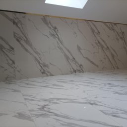 Artur Janoś Usługi ogólnobudowlane - Wnętrze wykończone białymi płytami imitującymi marmur, z delikatną złotą listwą przy suficie i oknem dachowym.
