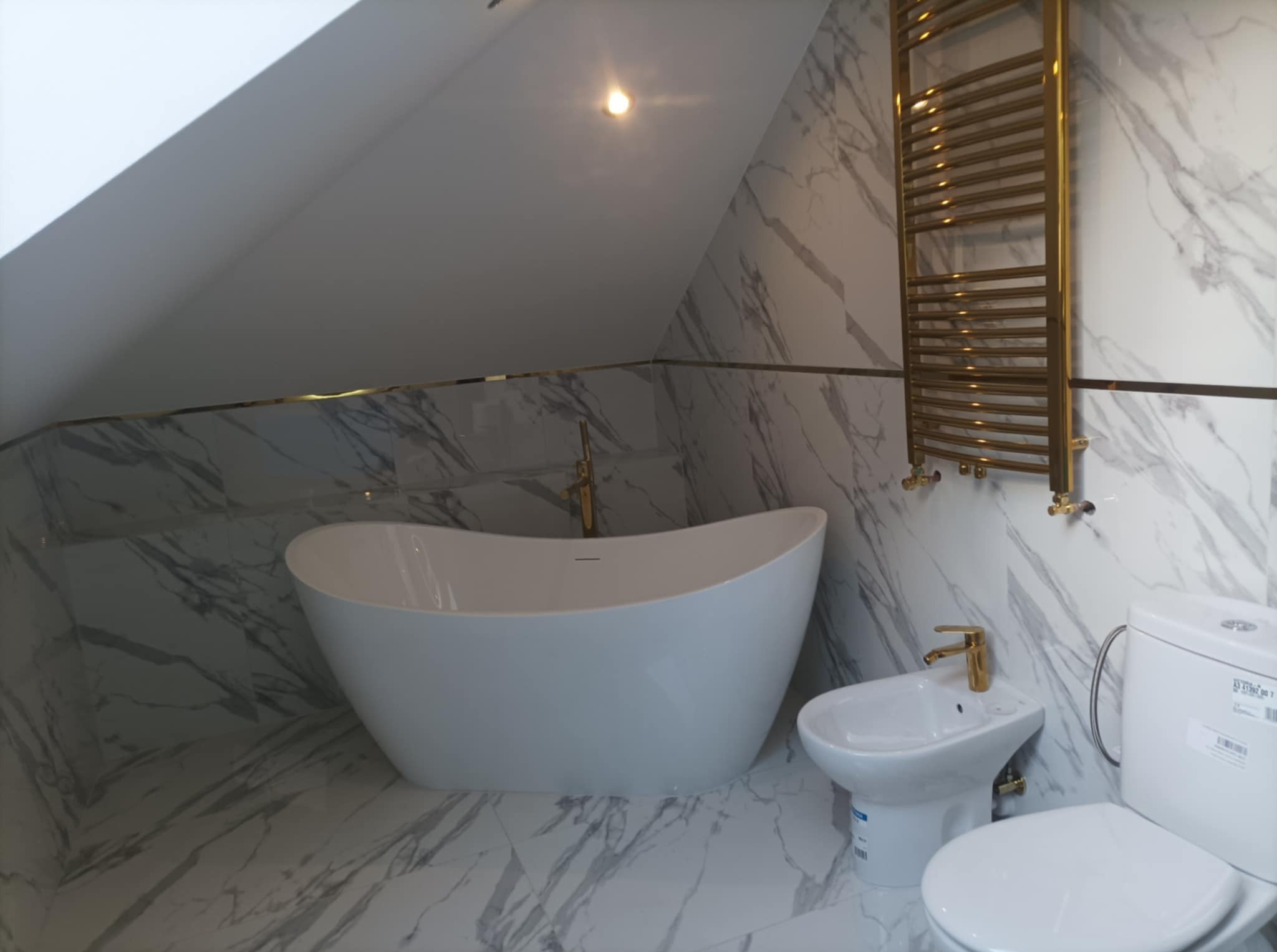 Nowoczesna łazienka na poddaszu z marmurowymi płytkami, wolnostojącą białą wanną, bidetem i toaletą w komplecie z bateriami w kolorze złotym oraz złotym grzejnikiem drabinkowym.