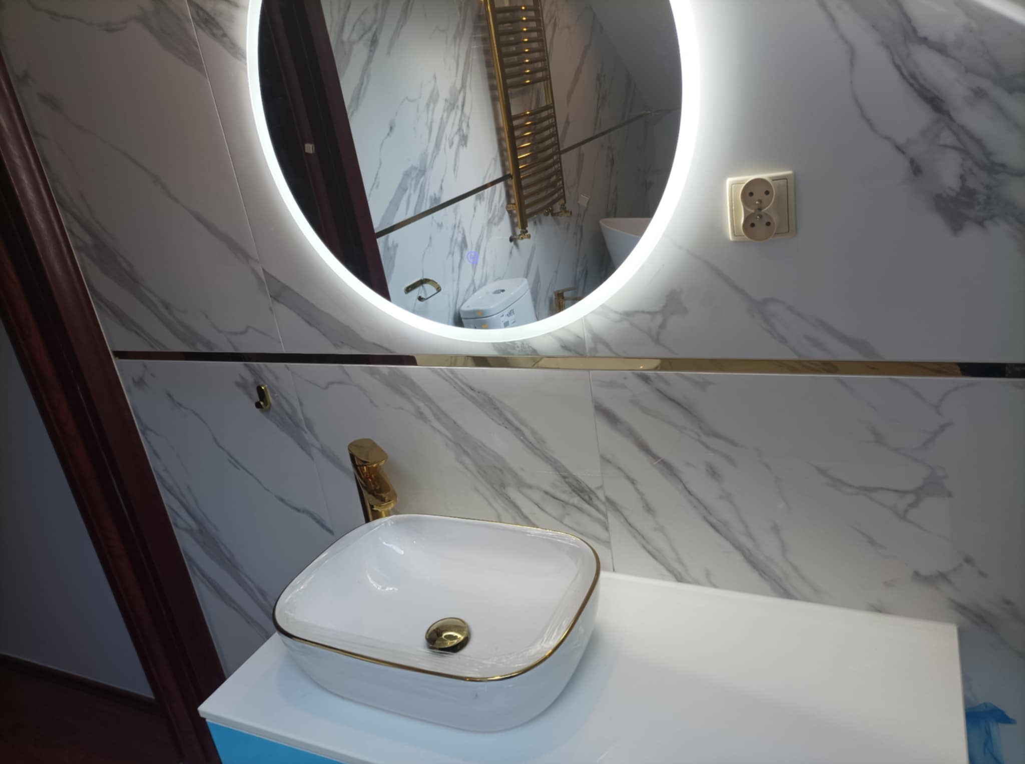 Nowoczesna łazienka z umywalką nablatową w białej, kwadratowej misie z złotym rantem, złotą baterią, okrągłym lustrem z podświetleniem LED, marmurową okładziną ścian i grzejnikiem drabinkowym...