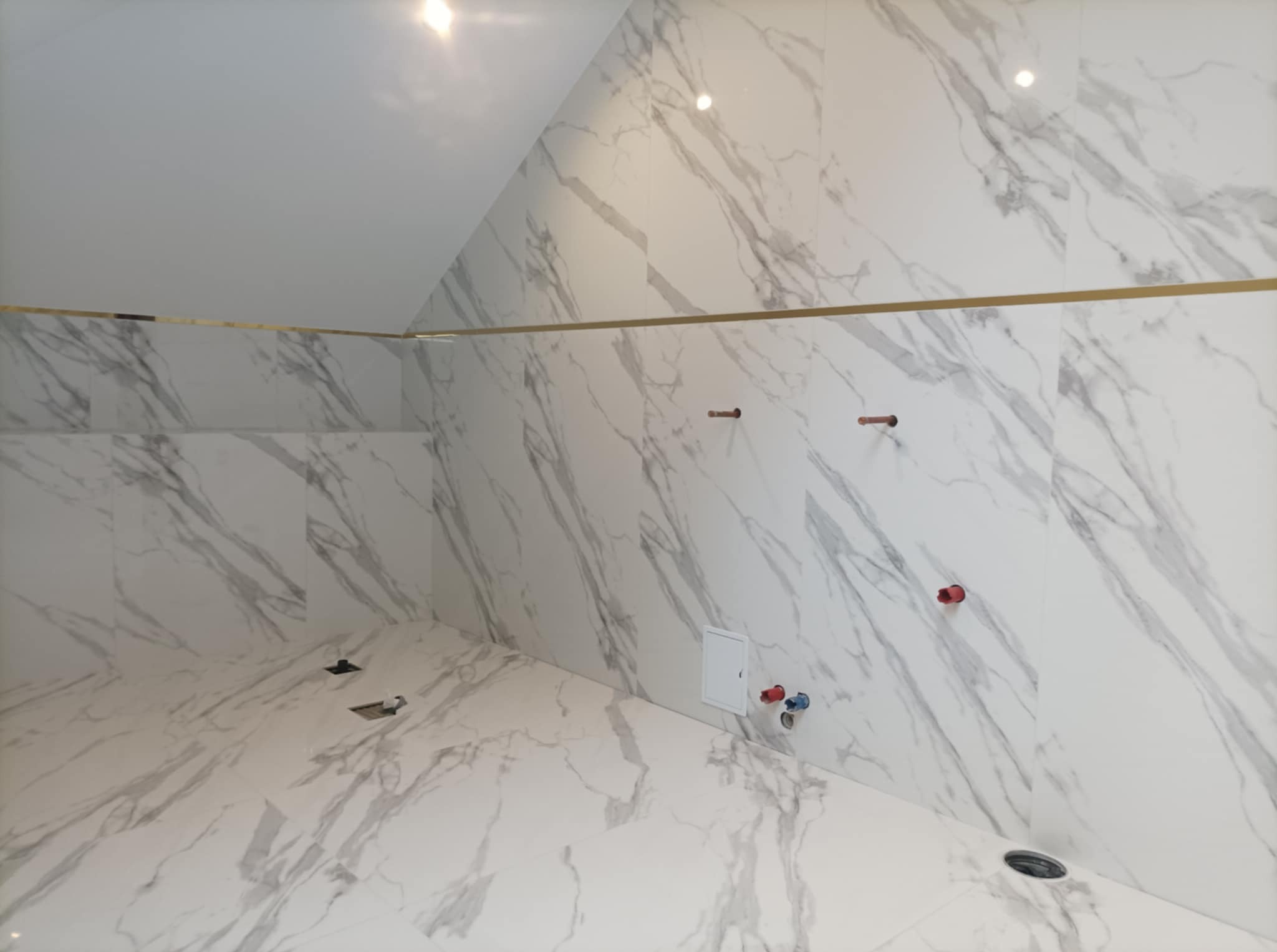 Ściany i podłoga w łazience wyłożone białymi płytkami imitującymi marmur, z widocznymi przyłączami hydraulicznymi i elektrycznymi przed montażem armatury, podkreślone złotym paskiem dekoracyjnym.