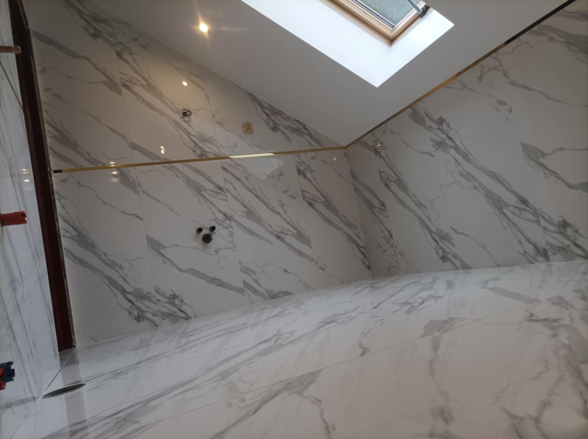 Wykończona łazienka na poddaszu ze skośnym sufitem, marmurowe płytki na ścianach i podłodze z złotymi listwami dekoracyjnymi, okno dachowe w suficie.